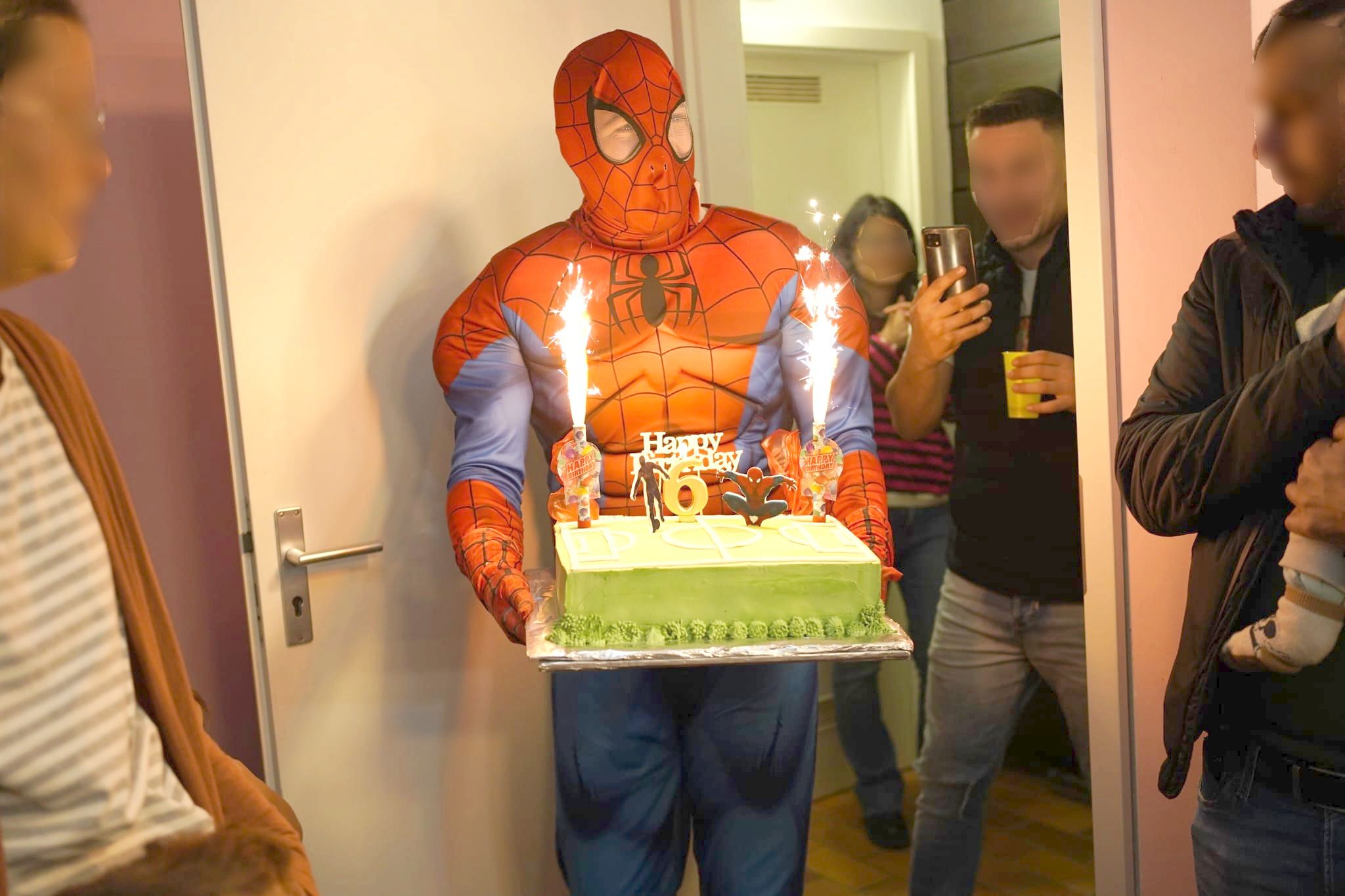 Auf dem Bild ist ein Animatuer als Spiderman verkleidet, der mit einer Torte in den Händen gerade ins Zimmertritt. Im Hintergrund sind Gäste zu sehen, die den Event fotografieren mit ihrem Mobilphone.